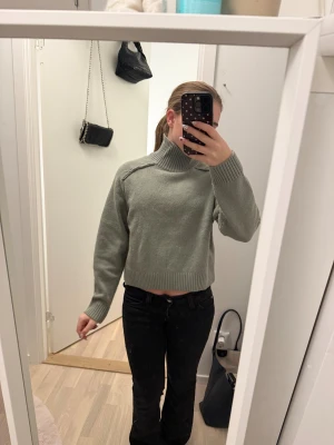 Grå croppad polotröja med ribbad krage - Snygg grå polotröja med croppad passform och ribbad hög krage. Tröjan har långa ärmar och breda ribbade muddar vid ärmslut och nederkant. Stickad i mjukt material, perfekt för lager på lager under höst och vinter. Enkel och stilren design som funkar till jeans eller kjol.