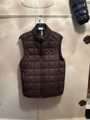 Brun quiltad dunväst från Replay - Säljer en brun quiltad dunväst från Replay med hög krage och dragkedja framtill. Västen har ett diskret märke på bröstet och är perfekt att slänga över en hoodie eller tröja. Fodrad och riktigt skön när det är kyligt ute.
