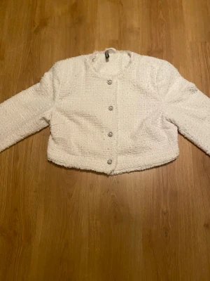 Vit kofta  - Säljer en vit kofta i tweed-liknande material med chanelldesign och snygga pärlknappar framtill. Koftan har rund halsringning, lång ärm och en croppad passform. 