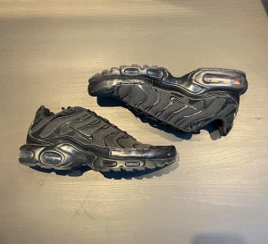 Nike Air Max Plus TN Svarta sneakers - Nike Air Max Plus TN sneakers i helsvart färg med mesh och syntetpaneler, ikoniska vågiga linjer och synlig Air Max-dämpning i sulan. Skorna har snörning, rund tå och platt sula. Perfekt för dig som gillar streetwear och sportig stil.