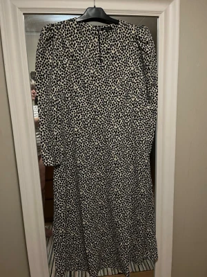 Prickig långklänning från Amelie & Me - Långklänning med svartprickigt mönster på beige botten från Amelie & Me. Klänningen har lång ärm med dragkedja vid ärmslut, rund halsringning och rak passform. Perfekt för dig som gillar statement-mönster och stilren look.