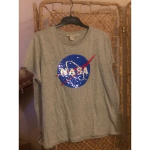Grå NASA t-shirt med paljetter - Grå t-shirt med rund hals och korta ärmar. På bröstet finns en stor paljettapplikation som kan vändas mellan NASA-loggan i blått, vitt och rött och en rymdfärja i vitt och svart. T-shirten är i mjuk bomull och har en avslappnad passform. Perfekt för dig som gillar rymden och coola detaljer.
