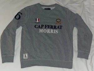 Grå Morris Cap Ferrat Storlek S - Grå tröja från Morris i mjuk bomull med broderade detaljer. Tröjan har rund hals, ribbade muddar och lång ärm med siffran 6 på höger arm. Storlek S. Köpt på NK för någon tusenlapp.