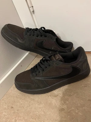 Nike Air Jordan 1 Low Travis Scott - Snygga Nike Air Jordan 1 Low Travis Scott i mörkbrun och svart mocka med svarta detaljer och vit kontrastsöm. Skorna har klassisk snörning, rund tå och ikoniska loggor på hälen. Perfekta för dig som gillar streetwear och unika sneakers.