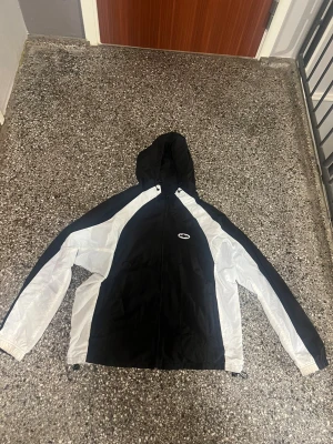 Corteiz regnjacka/vindjacka  - Snygg svart och vit vindjacka från Craft med huva och dragkedja framtill. Jackan har sportig design med vita paneler längs ärmarna och sidorna, samt Craft-logga på bröstet. Perfekt för dig som gillar streetwear och sportig stil.