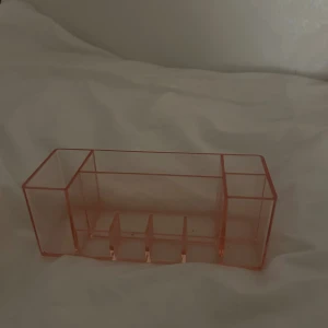 Rosa sminkförvaring  - Snygg och praktisk sminkförvaring i rosa transparent akryl med flera fack för borstar, pennor och småsaker. Perfekt för att organisera sminkbordet och hålla ordning på dina favoriter. Stilren design som passar in i de flesta rum.