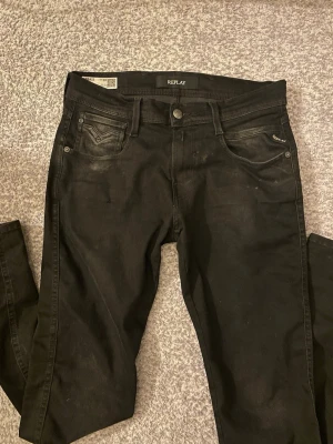 Svarta Replay Anbass jeans slim fit - Svarta jeans från Replay, modell Anbass, med slim fit passform. Klassisk femficksdesign, snygga detaljer på bakfickorna och bälteshällor. Jeansen är tillverkade i stretchigt jeansmaterial för en bekväm känsla och sitter tajt längs benen.