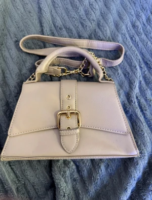 Beige handväska med kedja och bälte - Snygg beige handväska med guldfärgad kedja och dekorativt bälte framtill. Väskan har både handtag och axelrem, vilket gör den enkel att bära på olika sätt. Stilren design med detaljer i guld som ger en lyxig känsla. Perfekt för dig som gillar klassiska accessoarer.