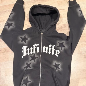 Svart hoodie med stjärnor och tryck - Svart hoodie med dragkedja och huva från Fortnite. Tröjan har stora vita bokstäver framtill och är täckt av svarta stjärnor med airbrush-effekt både fram och bak. Mjuk insida och känguruficka framtill. Perfekt för dig som gillar streetstyle och gaming.