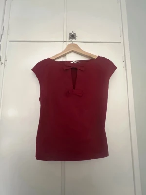 Röd blus perfekt för julen - Snygg röd blus med korta ärmar och en liten rosett framtill vid halsringningen. Perfekt nu till julen eller bara att ha vardagligt. Jättefin, passar med allt!