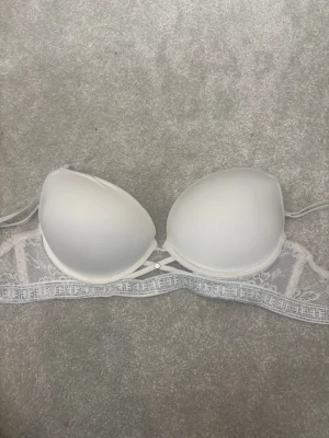 Vit bh med spets och strass från Censored - Snygg vit bh från Censored med vadderade kupor, spetsdetaljer och glittrande strass längs bandet. Justerbara axelband och dekorativt metallknapp framtill. Perfekt för dig som gillar lite extra bling och stil. 
