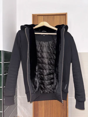 Moose Knuckles XL barn storlek  - Svart hoodiejacka från Moose Knuckles i barn storlek XL. Jackan har quiltad insida med glansigt mönster, mjuk fuskpäls längs öppningen och huva. Snyggt metallmärke på ärmen och grov dragkedja framtill. Perfekt för dig som gillar streetwear med exklusiv känsla.