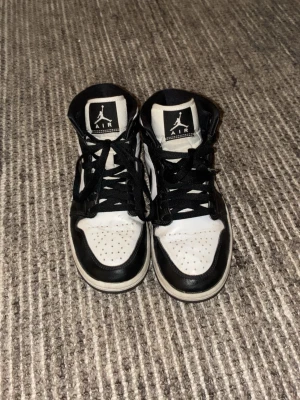Nike Air Jordan 1 svart/vit 37.5 - Nike Air Jordan 1 sneakers i klassisk svart och vit färg. High-top modell med snörning, rund tå och platt sula. Ikonisk Jumpman-logga på plösen och Air Jordan-märke på sidan. Tillverkade i skinn med syntetdetaljer. Perfekt för dig som gillar streetstyle och sneakers med attityd.