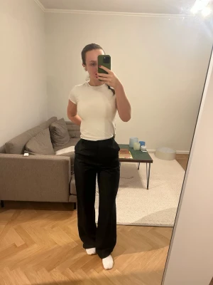 Svarta kostymbyxor med wide fit - Säljer ett par svarta kostymbyxor med hög midja och vida ben. Byxorna har klassisk look med pressveck och stängs med dragkedja och knapp. Perfekta för en clean och stilren outfit. Materialet känns mjukt och följsamt.