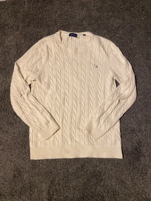 Vit kabelstickad tröja från GANT - Vit kabelstickad tröja från GANT i storlek S. Klassisk rund halsringning, ribbade muddar och broderad GANT-logga på bröstet. Tröjan är i mjuk bomull och har en tidlös preppy stil som funkar året runt.