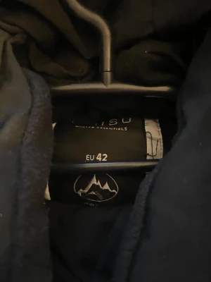 Svart dunväst med huva från Everest - Svart dunväst från Everest med broderad logga på bröstet. Västen har en stor, justerbar huva, två sidofickor och stängs med dragkedja och tryckknappar. Materialet är slitstark polyester och den quiltade designen ger extra värme. Perfekt för kalla dagar.