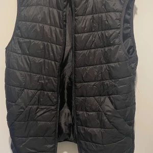 Svart quiltad dunväst från Zara - Svart quiltad dunväst från Zara i storlek S. Västen har en stilren look med horisontella sömmar, dragkedja framtill och två fickor. Perfekt att slänga över en hoodie eller tröja för extra värme. Lätt och smidig att bära.