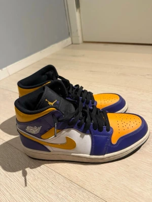 Nike Air Jordan 1 High Lakers - Nike Air Jordan 1 High sneakers med Lakers-inspirerad färgway. Skorna har lila och gula paneler i skinn, vit sidopanel och gul swoosh. Svart plös med gul Jumpman-logga, svart snörning och perforerad tå. Klassisk Air Jordan Wings-logga på sidan och platt svart sula.