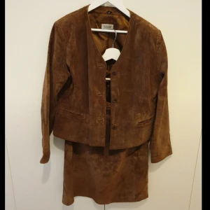 Brun mockakostym med kjol och kavaj - Snygg brun mockakostym från Jofama med kjol och kavaj. Kavajen har knappar framtill, två fickor och är helfodrad. Kjolen är rak och har dragkedja i sidan. 