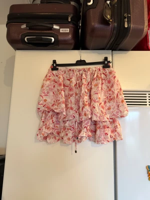 Odd Molly blommig volangkjol rosa - Supersöt kort kjol från Odd Molly med blommigt mönster i rosa, rött och orange. Kjolen har flera volanger, snörning i midjan och är gjord i ett tunt, luftigt material. Perfekt för dig som gillar boho-stil och vill ha något färgglatt och lekfullt. Storlek 2 som enligt deras storleksguide motsvarar en M. Mycket fint skick, jag hittar inga synliga anmärkningar eller defekter. 