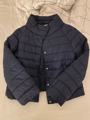 Mörkblå pufferjacka från Gina Tricot - Säljer en mörkblå pufferjacka från Gina Tricot i storlek 170. Jackan har quiltad design, hög krage och tryckknappar framtill. Perfekt till våren! Använd fåtal gånger! Köpt för ungefär 250kr!