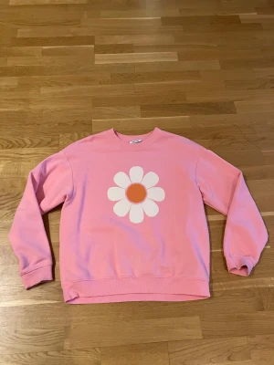 Rosa sweatshirt med blomtryck från Monki - Säljer en rosa sweatshirt från Moise med ett stort vitt och orange blomtryck på bröstet. Tröjan har rund hals, långa ärmar och ribbade muddar. Perfekt för dig som gillar färgglada och lekfulla plagg!