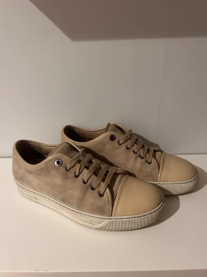 Beige sneakers från Lanvin i mocka - Snygga beige sneakers från Lanvin med ovandel i mocka och tåparti i slät skinn. Klassisk rund tå och platt sula med snörning framtill. Diskret design med ton-i-ton detaljer och vita kanter på sulan. Perfekt för dig som gillar stilrena och exklusiva sneakers.