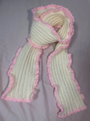 Söt och mysig handgjord halsduk🤍🎀 - Handvirkad halsduk i mjukt akrylgarn. Krämvit med dekorativ ljusrosa vågig kant. Ribbstruktur som gör den mjuk, varm och bekväm att bära. Unik och handgjord – finns endast ett exemplar. Ny och oanvänd. 144cm lång, 13cm bred.