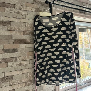 Grå  klänning med molnmönster Me&I - Supersöt grå långärmad från Me&I med vita moln över hela tyget.  lång ärm, rund halsringning och ett rosa knytband i midjan. Tillverkad i mjuk bomull som är skön att ha på sig. Perfekt för dig som gillar lekfulla prints och bekväma plagg.