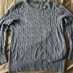 Ljusblå kabelstickad tröja från Polo Ralph Lauren - Säljer en ljusblå kabelstickad tröja från Polo Ralph Lauren med rund hals och långärmad design. Tröjan har ett litet rött broderat logomärke på bröstet och ribbade muddar vid ärmslut och nederkant. Perfekt för dig som gillar klassisk stil. Storlek S men passar nog M också