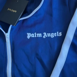 Blå track jacket från Palm Angels - Säljer en blå track jacket från Palm Angels med vita detaljer längs ärmarna och dragkedja framtill. Jackan har hög krage, vita ränder och logga på bröstet. Perfekt för en sportig och trendig look.