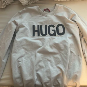Vit sweatshirt från HUGO - Säljer en stilren vit sweatshirt från HUGO med stor svart logga framtill. Tröjan har rund halsringning och långa ärmar, perfekt för en clean och avslappnad look. Tillverkad i mjuk bomull som är skön att bära hela dagen.