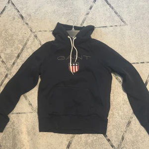 Marinblå hoodie från GANT - Säljer en marinblå hoodie från GANT med broderad logga och sköld på bröstet. Tröjan har huva med vita snören och en stor magficka. Perfekt för dig som gillar klassisk och sportig stil.