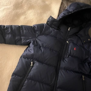 Mörkblå dunjacka från Polo Ralph Lauren - Mörkblå dunjacka från Polo Ralph Lauren med huva, tre dragkedjeförsedda fickor och klassisk röd logga på bröstet. Jackan är quiltad och har en snygg, sportig look med detaljer i metall och tryckknappar vid kragen. INGA DEFEKTER ‼️‼️‼️‼️‼️‼️‼️‼️ Storlek L barn, som en XS/S, köpt för 3500 