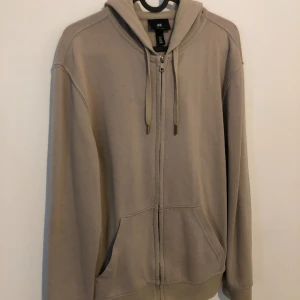 Beige hoodie med dragkedja från H&M - Snygg beige hoodie från H&M i regular fit med huva och snörning. Tröjan har dragkedja framtill och två stora fickor. Perfekt för chill dagar och enkel att matcha med jeans eller joggers. Mjuk bomullskänsla och långärmad design.
