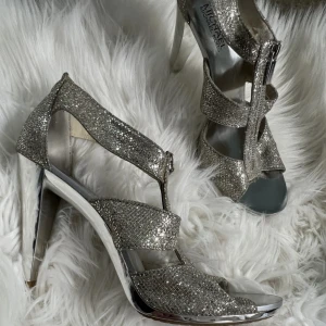 Glittriga silverklackar Michael Kors - Snygga klackar från Michael Kors i glittrigt silver med öppen tå och dragkedja framtill. Skorna har höga, blanka klackar och flera remmar i glittrigt syntetmaterial. Perfekta för dig som vill sticka ut med en glamorös look. Pris kan diskuteras 