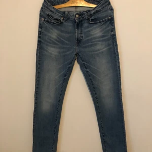 Slim fit jeans från Dressmann blå - Slim fit jeans från Dressmann i klassisk blå tvätt med snygga slitningar framtill. Modellen har fem fickor, knapp och dragkedja i midjan samt Dressmanns logga på insidan. Perfekt passform för dig som gillar smalare jeans.