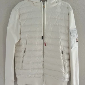 Vit pufferjacka från Moncler - Snygg vit pufferjacka från Moncler med huva och dragkedja framtill. Jackan har quiltad framsida, två fickor med dragkedja och en ikonisk Moncler-logga på ärmen. Materialmix med vadderad kropp och släta ärmar ger en clean och modern look. 10/10 skick, pris kan diskuteras.👍🏼