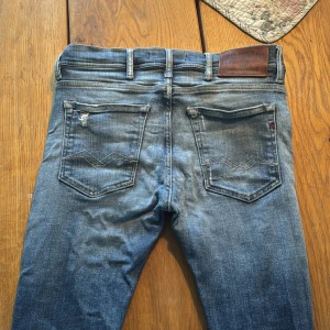 Replay blå slitna jeans, storlek 31/34 - Säljer ett par blå jeans från Replay med coola slitningar och lagade detaljer på benen. Klassisk femficksmodell med läderpatch bak i midjan och snygga sömmar på bakfickorna. Passar dig som gillar en avslappnad och edgy stil.