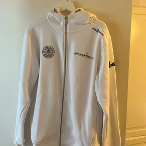 Vit hoodie Sail Racing Palma Vela oanvänd! - Vit hoodie från Sail Racing med dragkedja och huva. Framsidan har broderade loggor för Palma Vela och Real Club Nautico de Palma, samt detaljer på axel och ärm. Materialet är mjukt och känns som polyester, perfekt för dig som gillar snygga sportiga plagg.