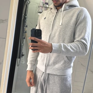 Grå hoodie från Polo Ralph Lauren - Snygg grå hoodie från Polo Ralph Lauren med klassisk blå logga på bröstet. Tröjan har huva med vit snörning, dragkedja framtill och två fickor. Insidan är mjukt fodrad i fleece för extra komfort. Perfekt för chill dagar och streetstyle. Passar S med 