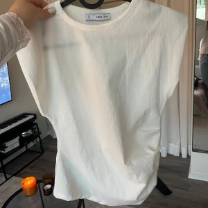 Vit topp från Mango - Säljer en helt ny aldrig använd vit topp från Mango i storlek S. Toppen är ärmlös med rund hals och har en lätt strukturerad yta som ger en snygg touch. Perfekt att matcha med jeans eller kjol för en clean look. Materialet känns mjukt och luftigt.