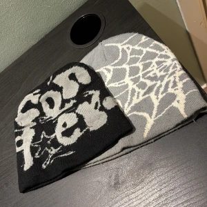 2 stickade mössor med tryck och spindelnät - Två coola stickade mössor, en svart med grått grafiskt texttryck och stjärnor, och en grå med vitt spindelnätsmönster. Perfekta för dig som vill sticka ut med edgy streetstyle. Mjukt material som håller dig varm under kalla dagar.