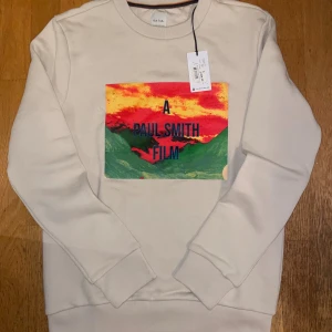 Krämfärgad Paul Smith limited edition sweatshirt - Snygg beige sweatshirt från Paul Smith med färgstarkt tryck på bröstet där det står 'A Paul Smith Film'. Tröjan är i 100% bomull och har rund hals, ribbade muddar och en cool biobiljett-detalj baktill. Riktigt skönt och tjockt material, oanvänd med prislapp kvar. 