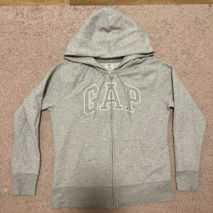 Grå hoodie med dragkedja från GAP - Snygg ljusgrå hoodie från GAP med dragkedja och stor logga framtill. Tröjan har huva med snörning, två fickor och är tillverkad i en mjuk bomulls- och polyestermix. Perfekt för en chill och avslappnad stil.