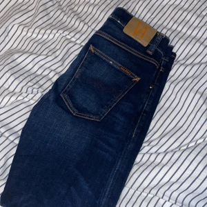 Mörkblå jeans från Nudie Jeans - Snygga mörkblå jeans från Nudie Jeans med klassisk femficksdesign och kontrastsömmar. Jeansen har en rak passform och är tillverkade i slitstarkt denim med läderpatch bak i midjan. Perfekta för dig som gillar stilrena och tidlösa jeans. Storlek W31 L30