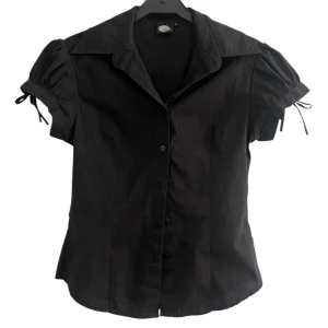 Svart kortärmad skjorta med snörning - Svart skjorta från ICE med kort puffärm och snörningsdetaljer vid ärmsluten. Skjortan har klassisk krage och knäppning framtill. Snygg passform.