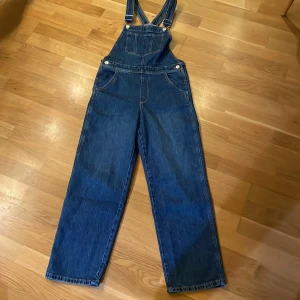 Blå denim hängselbyxor från H&M - Snygga blå hängselbyxor i klassisk jeansmodell från H&M. Byxorna har raka ben, justerbara axelband med metallspännen och flera fickor både fram och bak. Perfekt för dig som gillar en chill och retro vibe. Har använts en gång, säljer pg fel storlek.