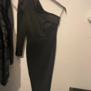 Svart one shoulder långklänning - Snygg svart långklänning med one shoulder-design och lång ärm på ena sidan. Klänningen har en tight passform och är perfekt för dig som vill sticka ut med en stilren och elegant look. Materialet känns mjukt och följsamt mot kroppen.