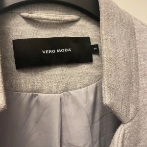 Ljusgrå kappa från Vero Moda - Stilren ljusgrå kappa från Vero Moda med rak passform och diskret krage. Jackan har två fickor framtill och stängs med en knapp. Tillverkad i mjukt material med slät insida, perfekt för dig som gillar minimalistisk look.
